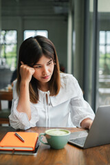 A young woman in a white blouse working on her laptop in a cozy cafe, with a notebook and a cup of coffee on the wooden table.