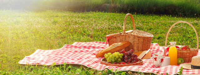 Picnic blanket with wicker basket, snacks and juice and on green grass during sunny day. Banner...