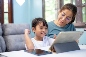 A young girl is sitting at a table with her mother, looking at a tablet.