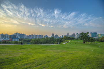 Olympic Park, Songpa-gu, Seoul, South Korea - June 19, 2021: Summer view of tourists on trail and lawn at Mongchontoseong Fortress against highrise apartments at sunset