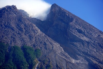 Mount Merapi in Yogyakarta, Indonesia, an active volcano with a split gray peak emitting white smoke, seen clearly against a bright blue sky