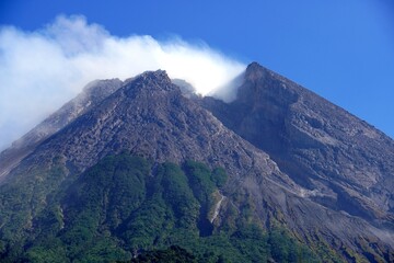 Fototapeta premium Mount Merapi in Yogyakarta, Indonesia, an active volcano with a split gray peak emitting white smoke, seen clearly against a bright blue sky