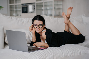 woman in glasses lies comfortably on a white couch, using a laptop. She is smiling and appears focused, creating a cozy and productive atmosphere