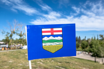 The Alberta provincial flag, featuring a blue field with a yellow shield depicting mountains, a river, and a cross, waves proudly in the wind against a backdrop of a clear blue sky and green landscape