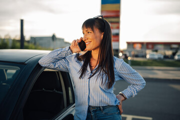 Fototapeta premium Young happy woman leaning on her car on parking lot and taking on phone