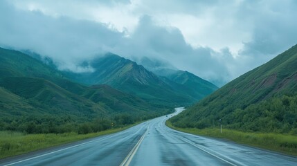 Naklejka premium A winding asphalt highway road leading towards green mountains under a cloudy morning sky