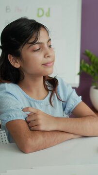 Vertical shot of Indian schoolgirl pronouncing sounds when sitting at table at logopedic session