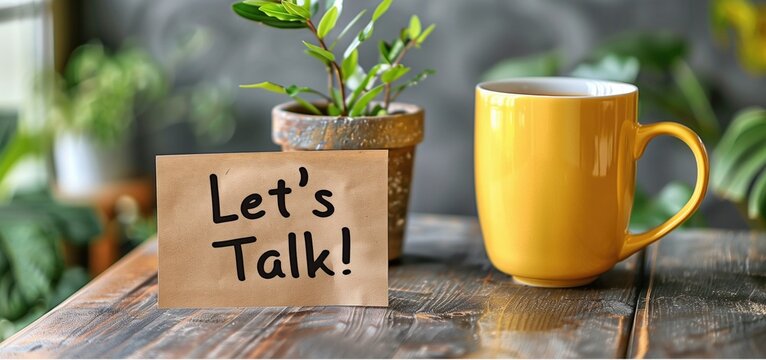 Brown paper note with "Let's Talk!" text next to potted plant and yellow mug on wooden table. Concepts of communication, conversation, and casual meeting settings.