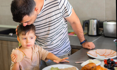 dad and son in modern kitchen eating breakfast.father doing tasty sandwich,smiling happy kid. boy waiting the meal.man kissing child,giving bread with ham.plates with food on counter.morning time