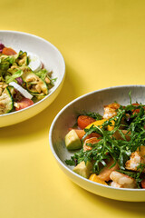 A fresh and vibrant salad featuring grilled shrimp, arugula, cherry tomatoes, avocado, and citrus segments, served in a modern bowl against a bright yellow background.