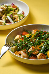 A fresh and vibrant salad featuring grilled shrimp, arugula, cherry tomatoes, avocado, and citrus segments, served in a modern bowl against a bright yellow background.