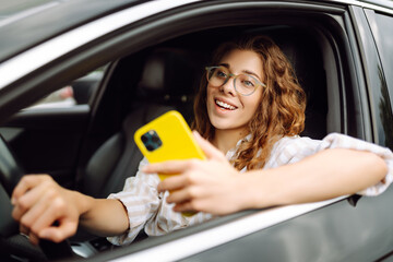 Young woman with curly hair using a phone while driving a car on a city street on a sunny day. Leisure, travel, technology. Carsharing.