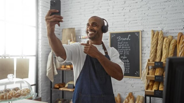 Young man in a bakery shop smiling and pointing at his phone while on a video call, surrounded by bread and pastries against a brick wall interior.