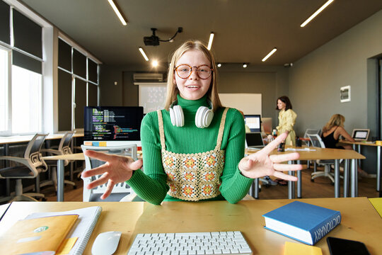 POV of young woman wearing glasses smiling at camera and gesturing while speaking via video chat in school classroom