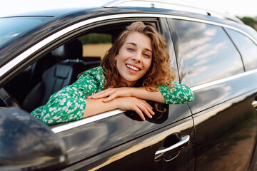 A young traveler woman enjoying a sunny day by the roadside in a green dress, leaning out of a car...