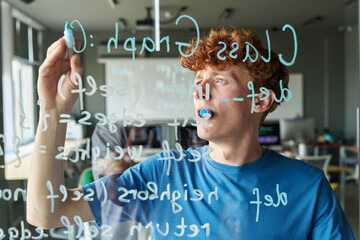 Student writing computer programming code on glass wall during IT class in school or college...