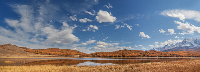 Autumn landscape with a lake, picturesque sky with white clouds, forested slopes and snow-covered peaks, panoramic view