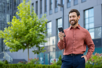 Smiling businessman standing outside in modern city environment using phone. Wearing stylish shirt and looking pleased while staying connected. Represents success, communication