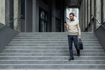 Confident businessman descending stairs holding phone and briefcase. Wearing glasses, sweater, and...