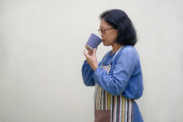 Asian woman standing side ways, wearing apron, sipping a cup of hot coffee