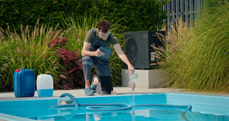 Pool technician pouring liquid into the pool while using a tablet, surrounded by chemical containers and greenery.