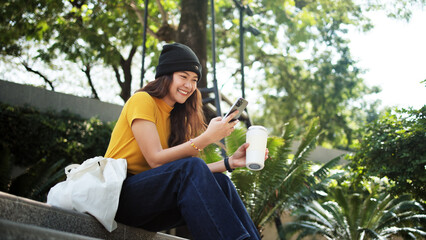 Gen Z eco go green woman asia sit happy relax at tree public park city life enjoy typing mobile...