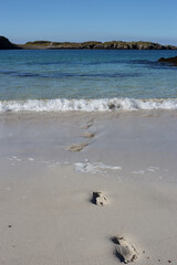Footprints on a sandy beach with small waves breaking onto the beach and rocks in the background.