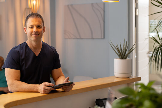 Holding tablet, male doctor, healthcare worker in scrubs smiling at reception in clinic, copy space