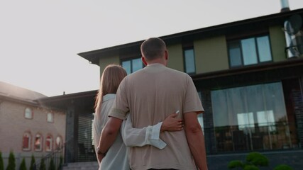 The couple is buying a big house. New large modern house. The family chooses a property, standing in front of building and discovering. Married couple looking at house, standing and hugging together