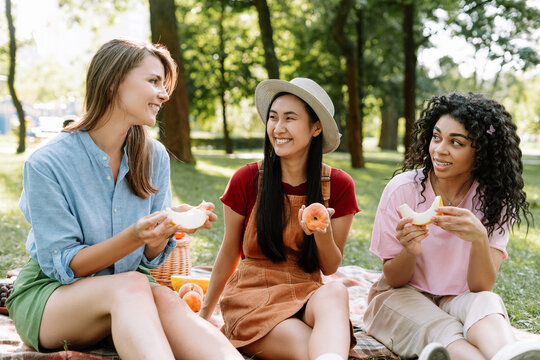 Smiling, multinational women, friends enjoying peach during picnic in a park, talking