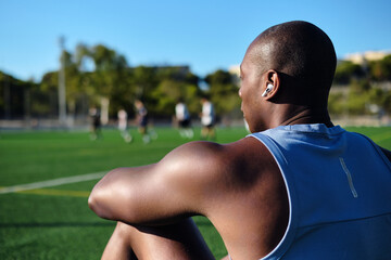 Young athletic man soccer player wear wireless earphones, blue athletic tank top sitting on grassy sports football field, resting after game or workout. Take break, sports, lifestyle © Alex Tihonov