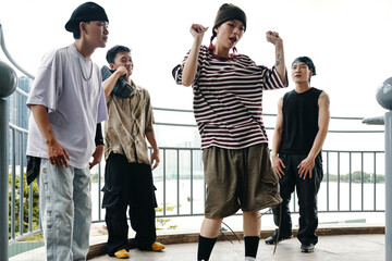 Group of four young Asian individuals practicing hip-hop dance moves on outdoor platform with cityscape visible in background exuding energy and enthusiasm for dance