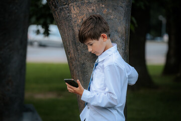 A young boy dressed in a white shirt and blue plaid tie leans against a tree while intently looking...