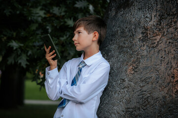 A young boy dressed in a white shirt and blue plaid tie leans against a tree while intently looking...