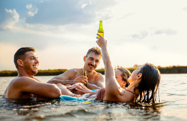 Group of young friends having fun and enjoying a summer day swimming with rubber tube and drinking beer at the lake.