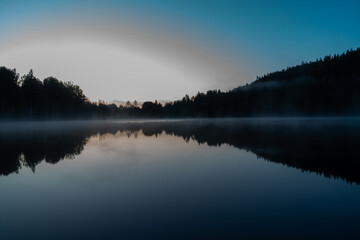 A tranquil lake reflects the landscape and sky during twilight, with silhouettes of trees lining the shorelines on either side. Mist hovers gently above the water's surface