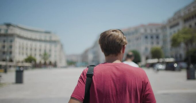 Young man exploring sunny city square, back view with a focus on his travel bag