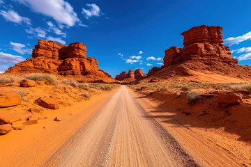 Selbstklebende Fototapeten An expansive desert landscape showcasing a long dirt road leading through stunning red rock formations under a deep blue sky, capturing the essence of open-road adventure. © DKPhoto
