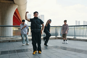 Group of dancers performing under bridge in urban setting, showcasing energetic and dynamic dance moves. Urban skyline visible in background
