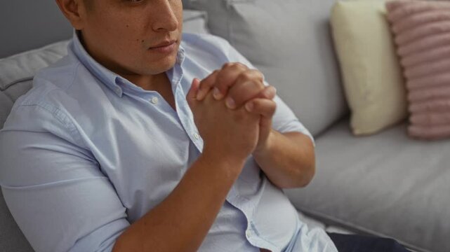 Young man praying indoors at home, sitting on a sofa in a living room