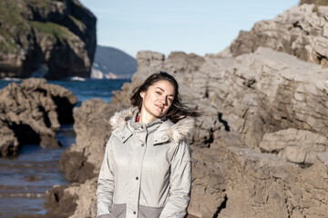 Naklejka premium A woman is standing on a rocky beach with her hair blowing in the wind. She is wearing a grey jacket and smiling