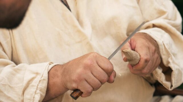 A man in ancient clothes is carving a wooden stick with a knife. Craft culture of the Baltics. Historical reenactment. Rumsiskes, Lithuania- 07 05 2024. 
