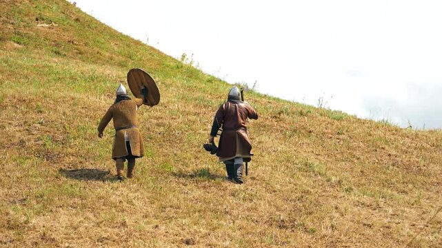Two men in medieval warrior clothes walk away. Raises hand with shield. Historical reenactment. Rumsiskes, Lithuania- 07 05 2024. 