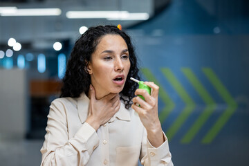 Hispanic businesswoman in office environment using throat spray for relief. Depicts concepts of...