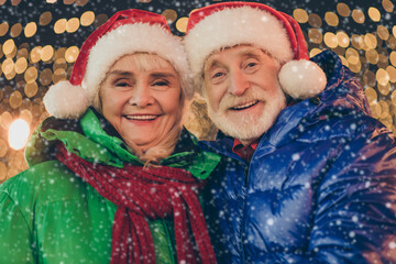 Two retired pensioner grey white hair people affectionate couple woman man celebrate x-mas christmas jolly event walk outside under illumination wear santa claus headwear