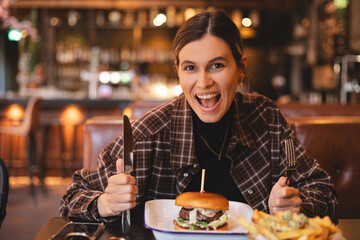 Woman eating a burger in a restaurant, knife and fork prepared. Cheat meal, fast food. Portrait of a young lovely hungry woman sitting in the fast junk food restaurant cafe. Unhealthy eating. Wow.