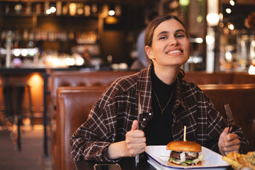 Woman eating a burger in a restaurant, knife and fork prepared. Cheat meal, fast food. Portrait of a young lovely hungry woman sitting in the fast junk food restaurant cafe. Unhealthy eating. 