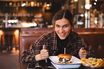 Woman eating a burger in a restaurant, knife and fork prepared. Cheat meal, fast food. Portrait of a young lovely hungry woman sitting in the fast junk food restaurant cafe. Unhealthy eating. 