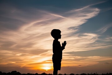 Fototapeta premium Silhouette of a Child in Prayer under a Stunning Sunset Sky - Embracing Light, Freedom, and Community