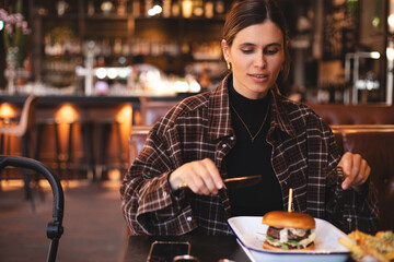 Woman eating a burger in a restaurant, knife and fork prepared. Cheat meal, fast food. Portrait of...
